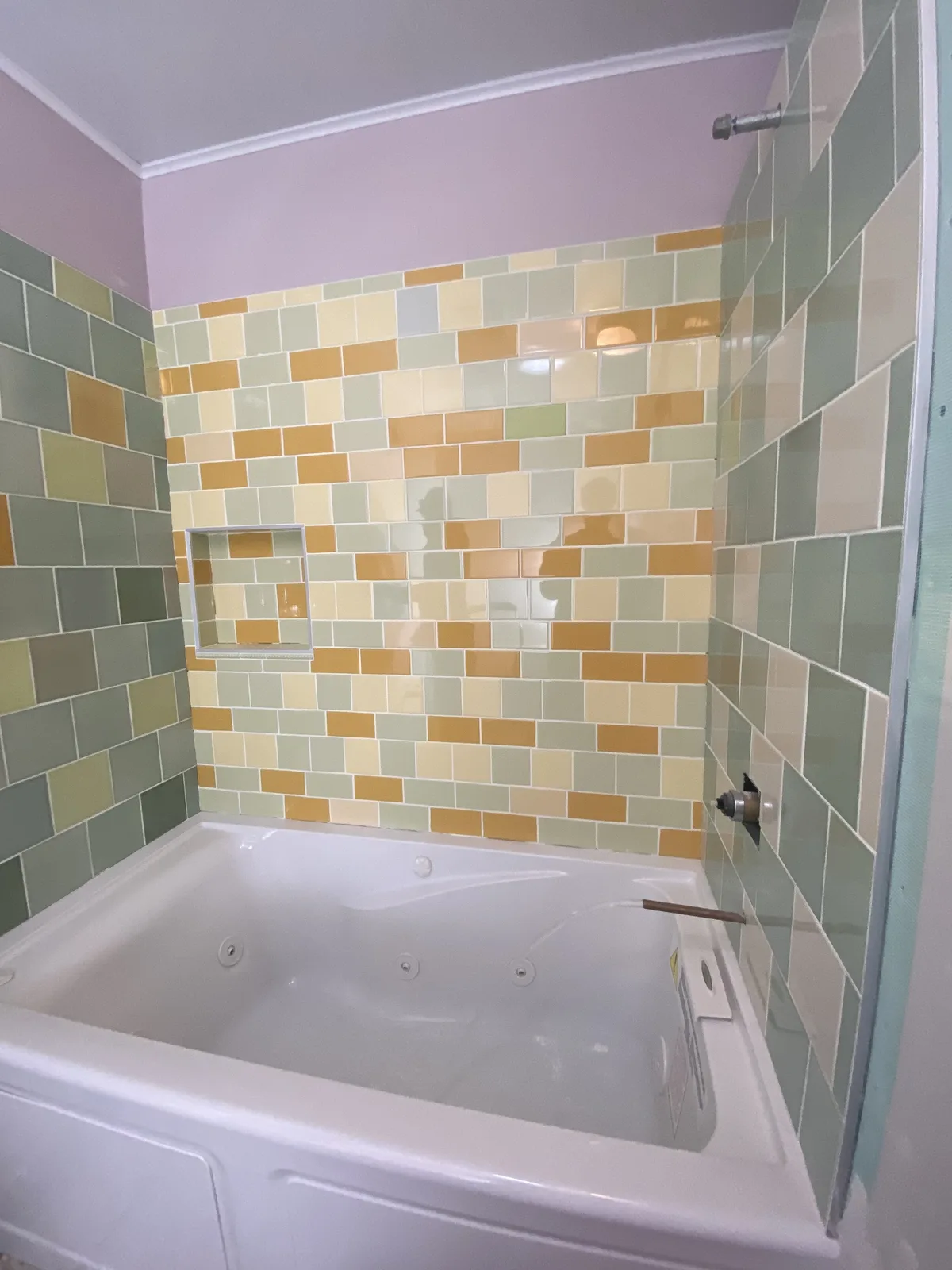 Jetted tub set in a surround of sage, butter yellow, and amber square tile with a framed niche, awaiting valve trim