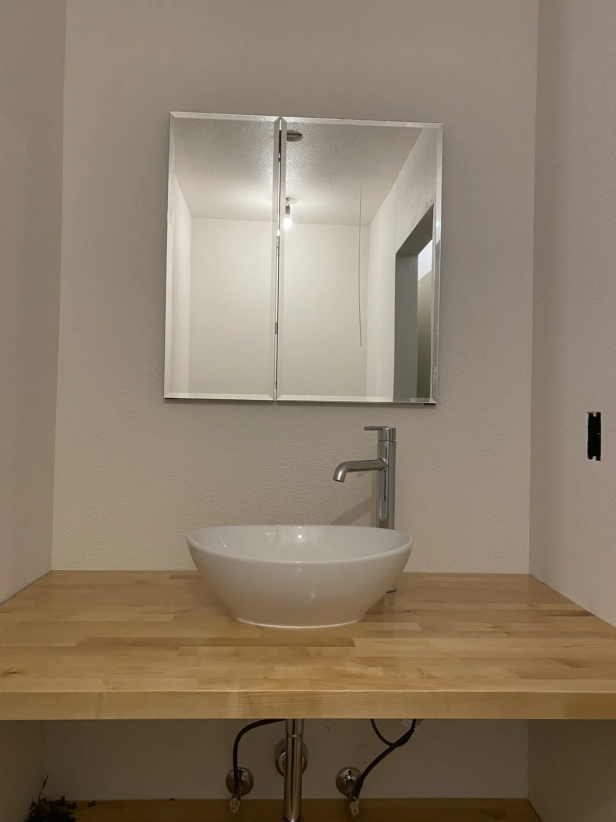 White vessel sink and tall chrome faucet on a butcher block counter below twin beveled mirrors